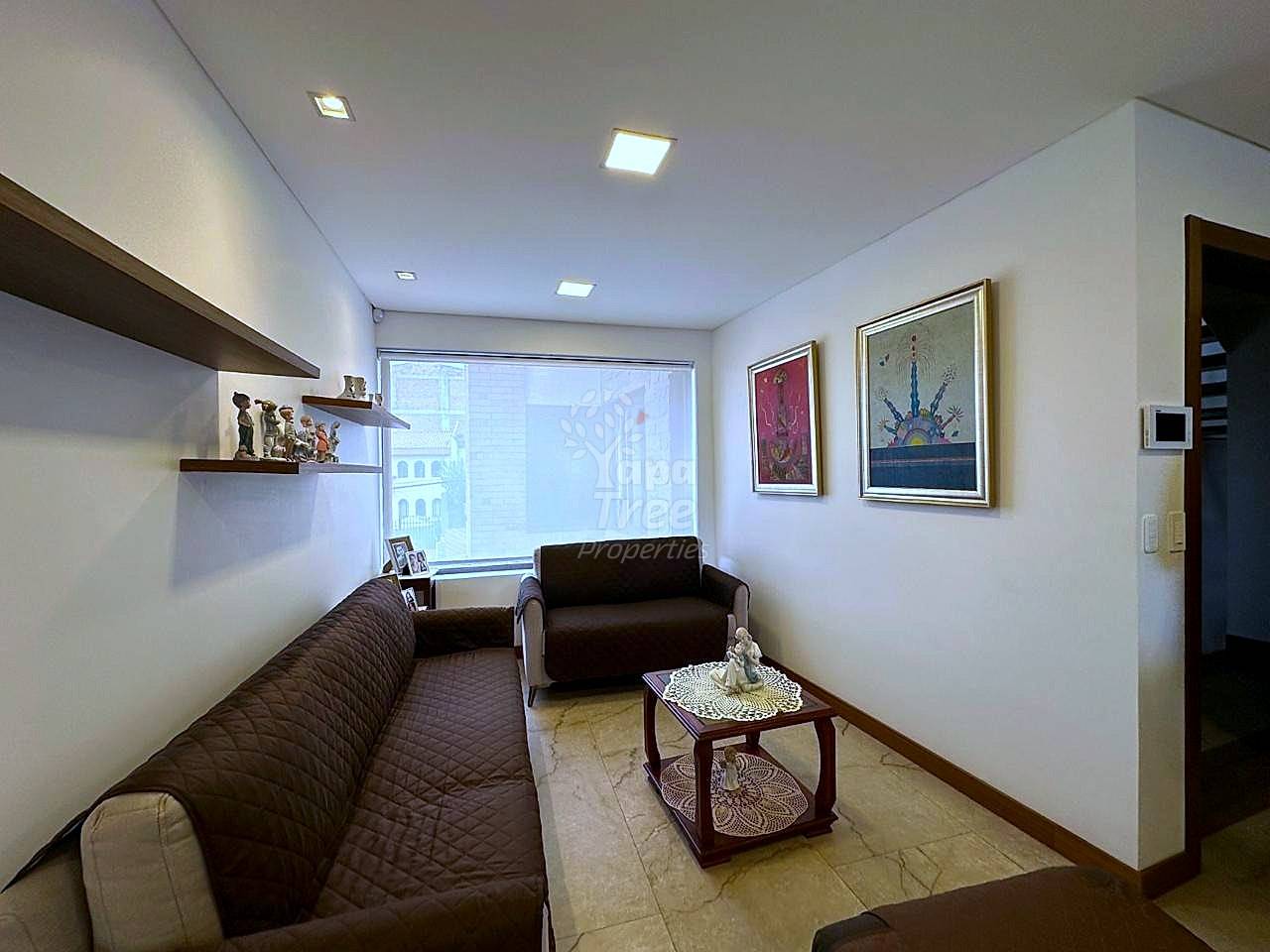 Living room with dark brown leather sofas, wooden coffee table, framed wall art, floating shelves, recessed ceiling lights, and large window with city view.