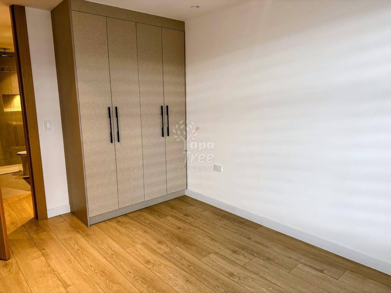 Bedroom with built-in four-door beige wardrobe, wooden plank flooring, white walls, and natural light from doorway.