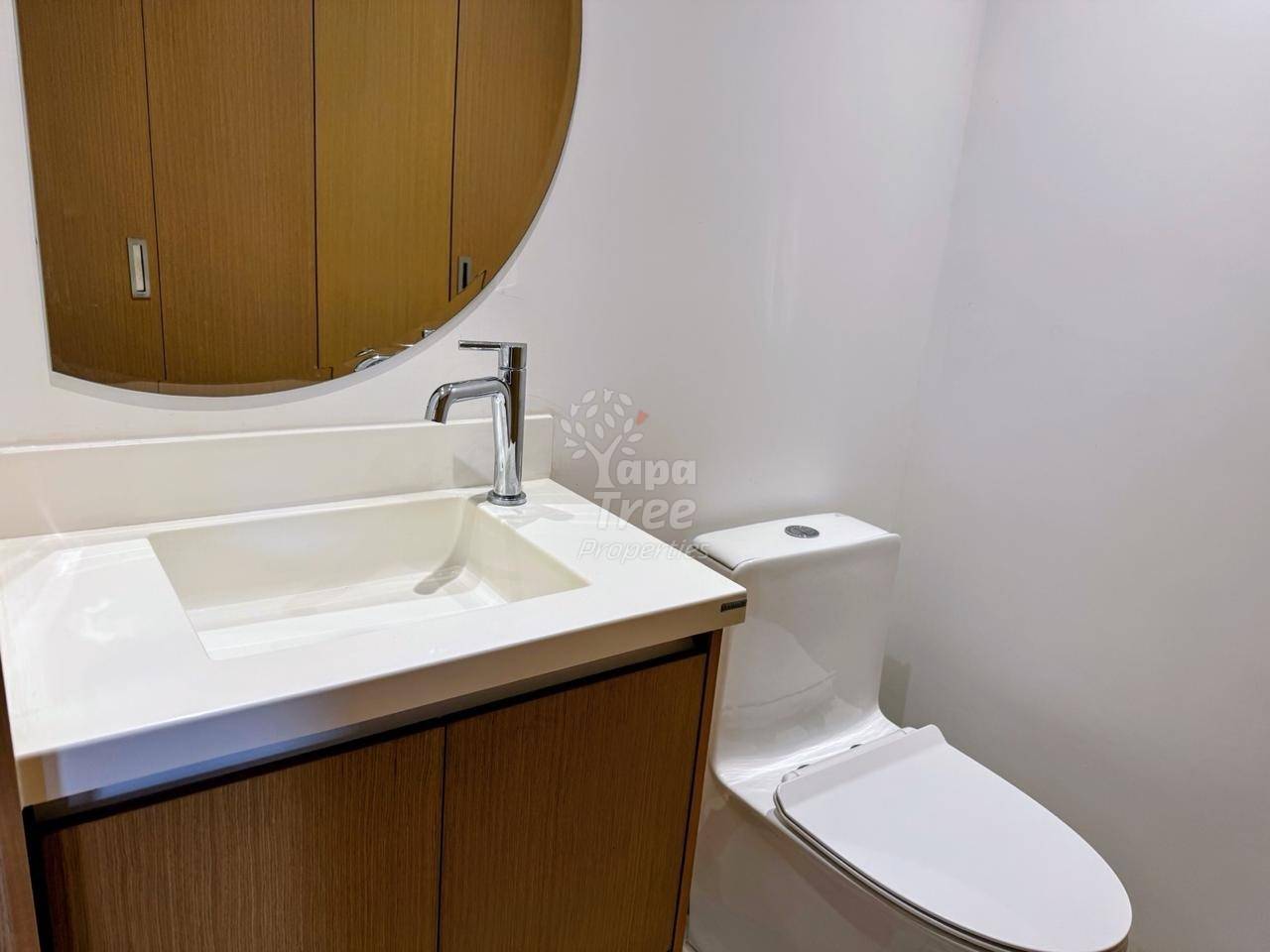 Bathroom with white walls, wood vanity cabinet, white countertop sink with chrome faucet, curved wood mirror cabinet, and white toilet.