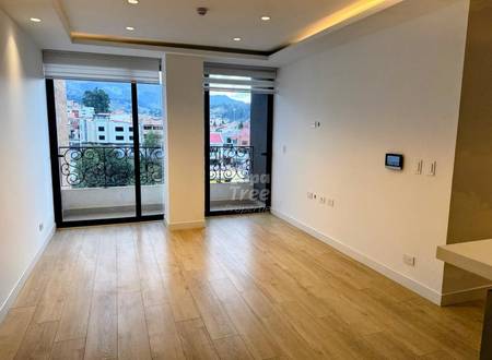 Living room with wood laminate flooring, cream walls, recessed ceiling lights, two black-framed glass doors opening to balconies overlooking Cuenca mountains and city buildings.