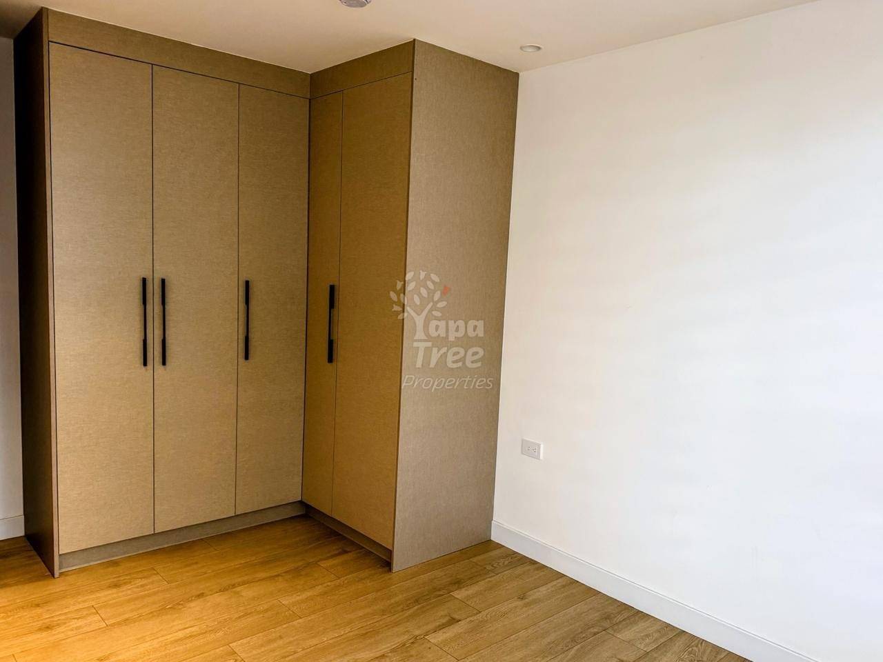 Bedroom corner with large built-in wooden wardrobe featuring four doors and black handles, light wood flooring, white walls, and recessed ceiling light.