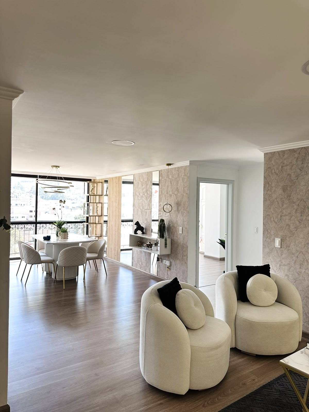 Open-concept living area with two cream-colored rounded armchairs, wooden flooring, patterned wallpaper, and dining table visible beyond.