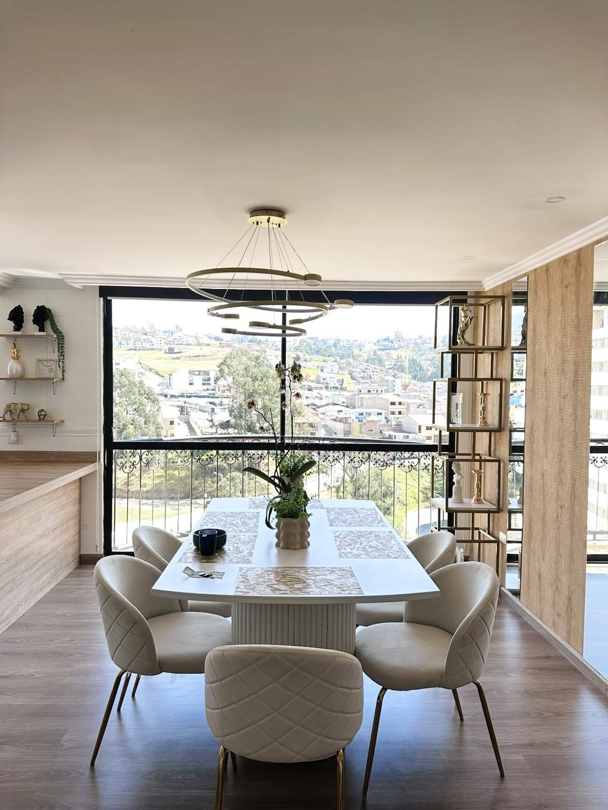 Dining room with white marble table, cream upholstered chairs, modern ring pendant light, and floor-to-ceiling windows overlooking Cuenca cityscape.