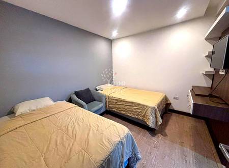 Bedroom with two single beds with beige bedding, gray accent chair, recessed ceiling lights, wood-look flooring, and floating wooden shelves on right wall.