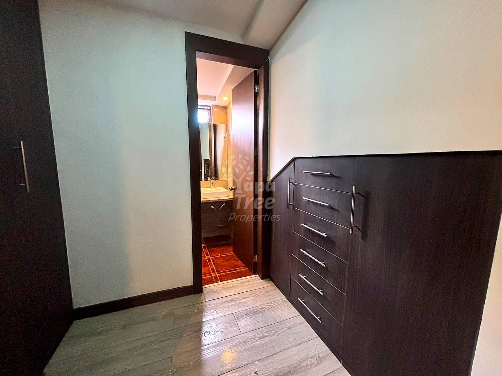 Bedroom hallway with gray tile flooring, dark wood dresser with five drawers, white walls, and open doorway to bathroom beyond.