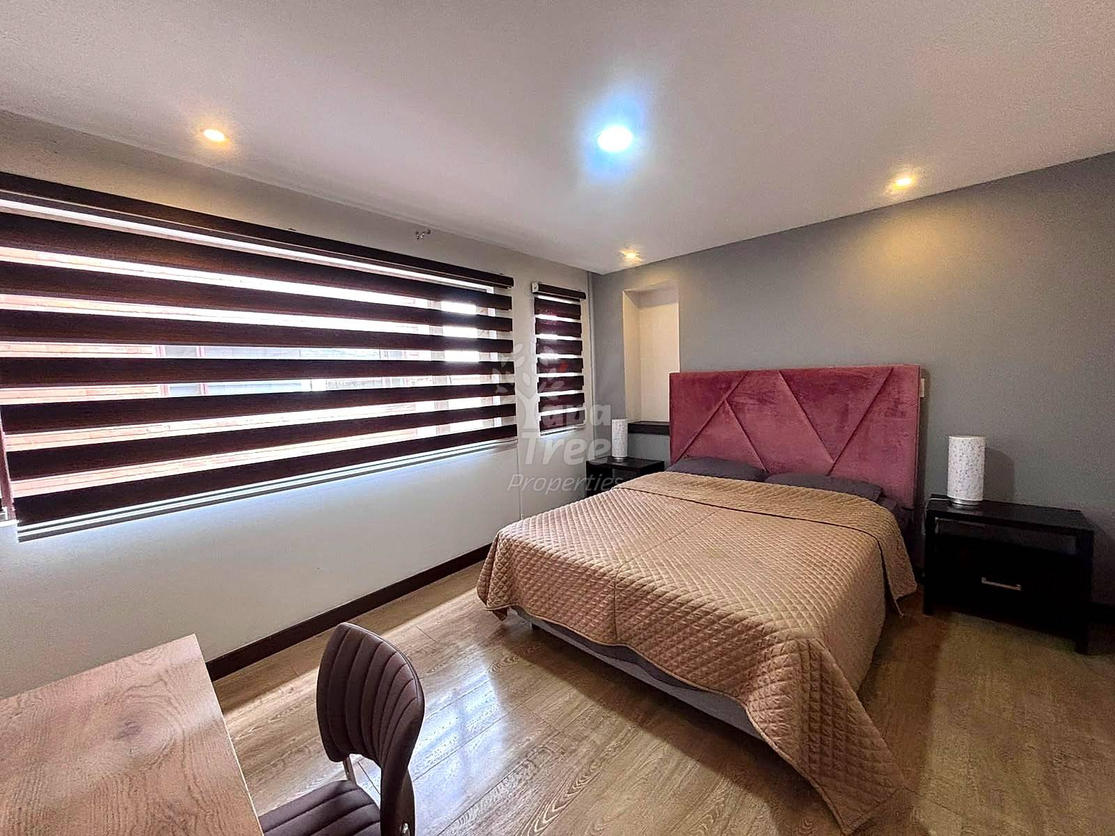 Bedroom with queen bed, burgundy upholstered headboard, wood nightstand, striped roller blinds, recessed ceiling lights, and wood-look flooring.