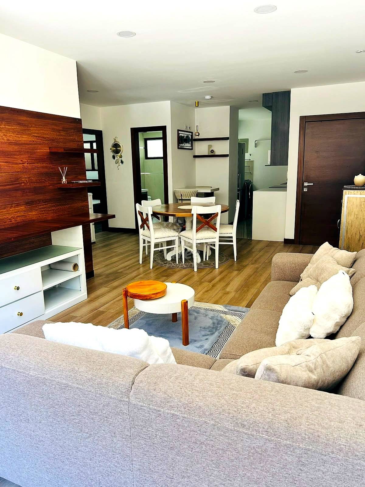 Open-concept departamento living area with beige sectional sofa, round white coffee table, wooden dining table with white chairs, and wood-paneled accent wall with recessed ceiling lights.