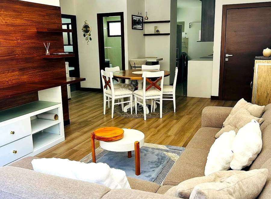 Open-concept departamento living area with beige sectional sofa, round white coffee table, wooden dining table with white chairs, and wood-paneled accent wall with recessed ceiling lights.