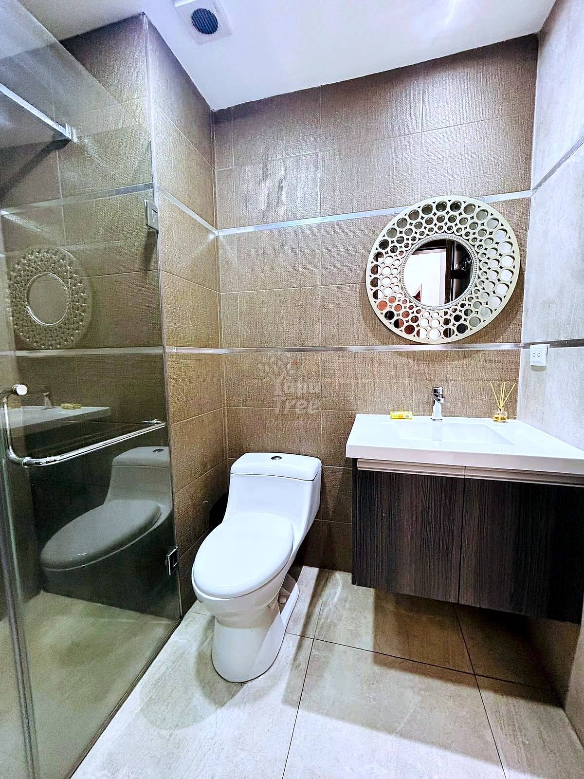 Bathroom with beige tile walls, white toilet, dark wood vanity with white sink, glass shower enclosure, and circular decorative mirror.
