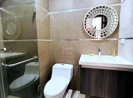 Bathroom with beige tile walls, white toilet, dark wood vanity with white sink, glass shower enclosure, and circular decorative mirror.