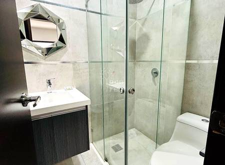 Modern bathroom with glass enclosed shower, white toilet, dark vanity cabinet, white sink, and geometric mirror on tiled walls.