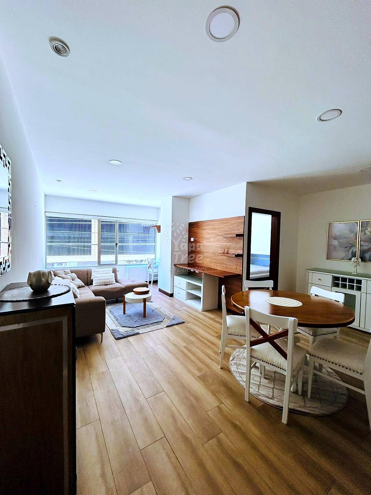 Open-concept living and dining area with wooden flooring, beige sofa, dining table with chairs, wood accent wall, recessed ceiling lights, and large windows.