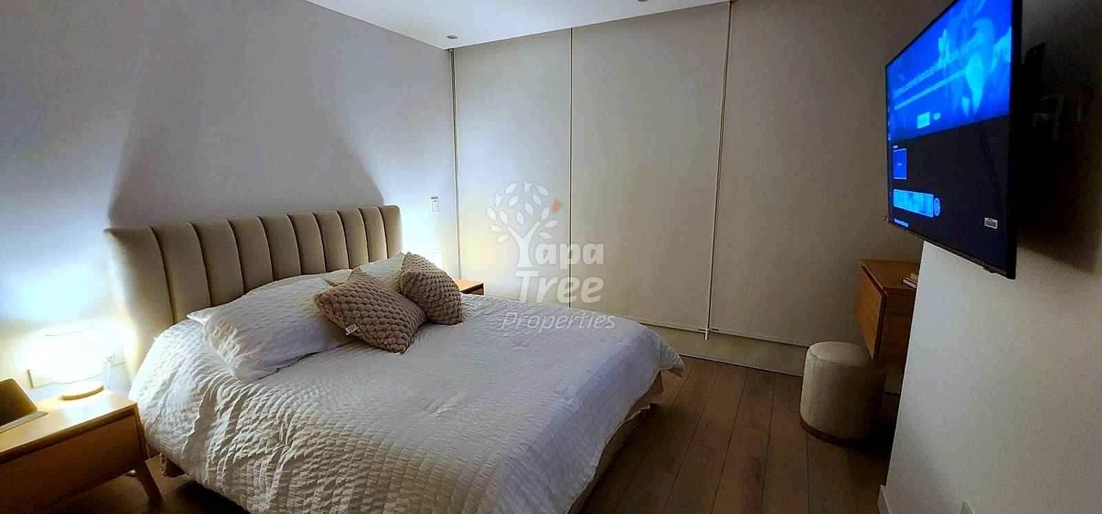 Bedroom with queen bed, beige upholstered headboard, bedside lamp, wall-mounted TV, wooden nightstand, and hardwood flooring.
