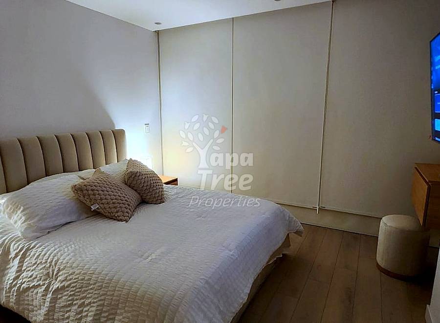 Bedroom with queen bed, beige upholstered headboard, bedside lamp, wall-mounted TV, wooden nightstand, and hardwood flooring.
