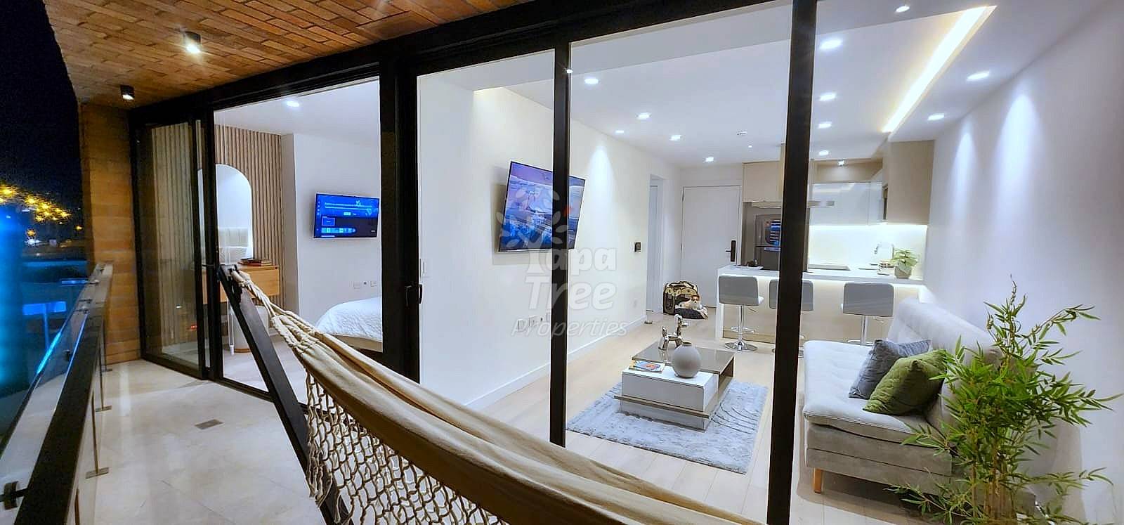 Open-plan departamento interior at night with black-framed glass partitions separating bedroom, living area, and cocina; wood ceiling, recessed lighting, modern furnishings visible.