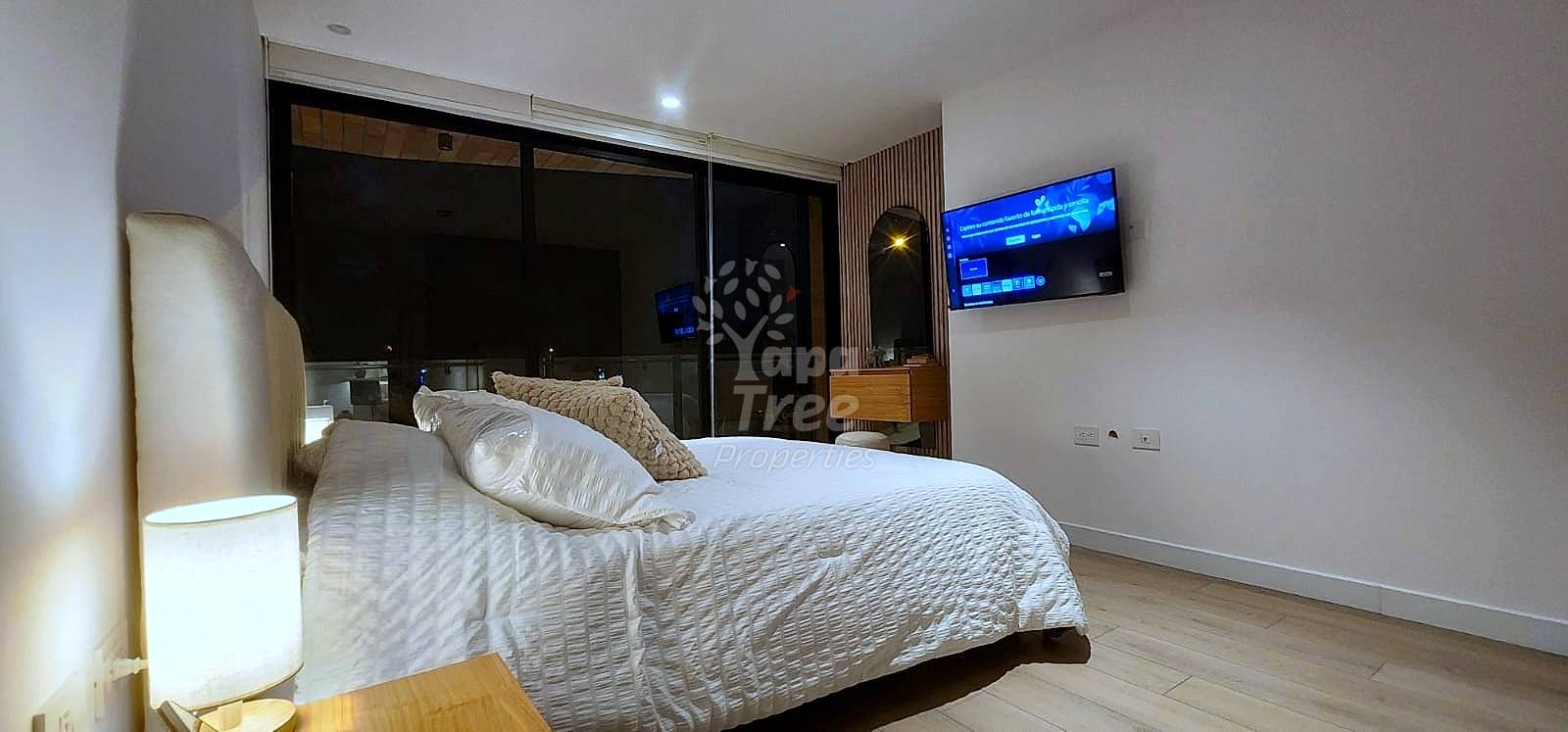 Bedroom with queen bed, white bedding, pillows, wall-mounted television, large black-framed windows, wooden nightstand with lamp, and light wood flooring.
