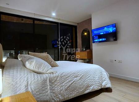 Bedroom with queen bed, white bedding, pillows, wall-mounted television, large black-framed windows, wooden nightstand with lamp, and light wood flooring.