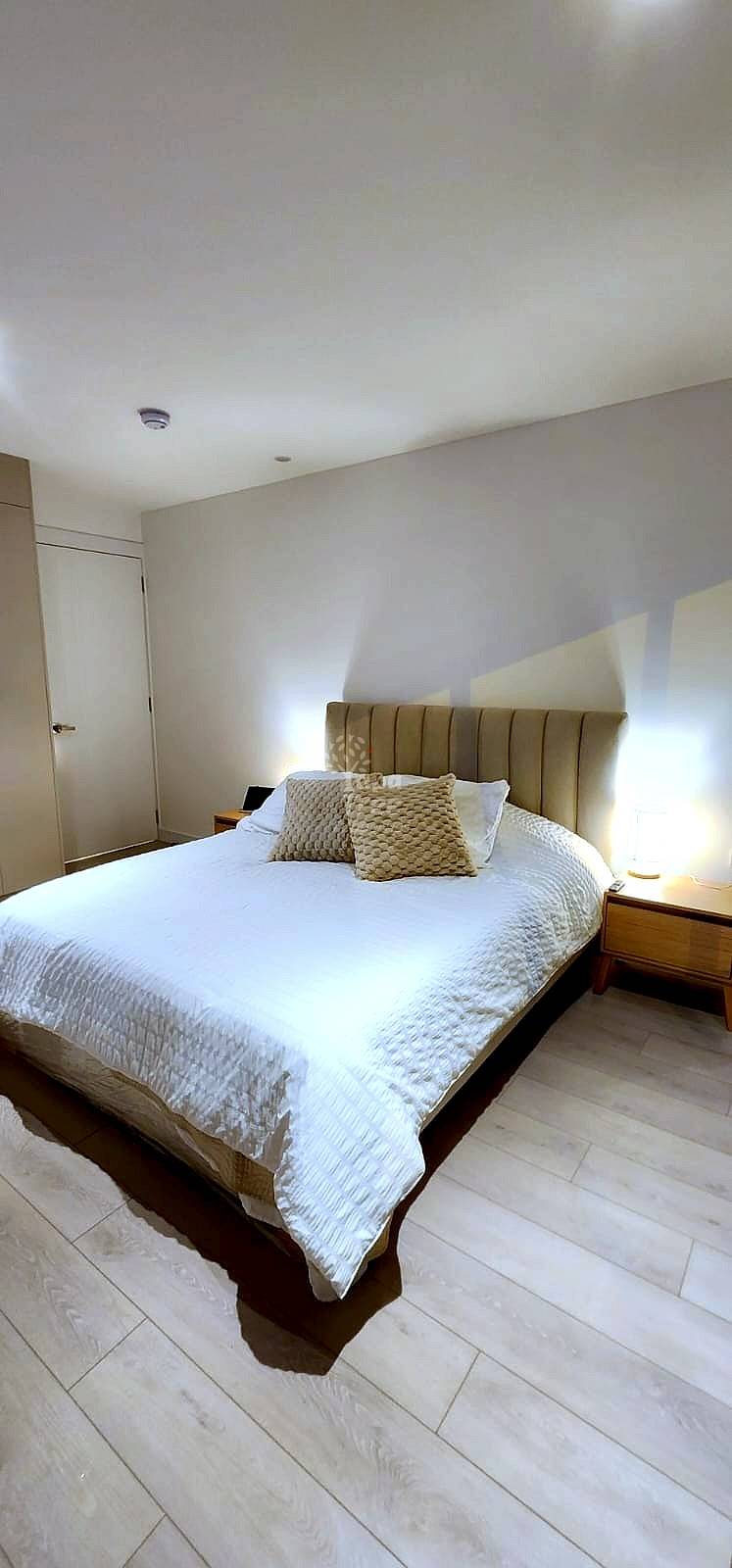 Bedroom with queen bed, upholstered headboard, white linens, two beige pillows, wooden nightstand with lamp, cream walls, and recessed lighting.