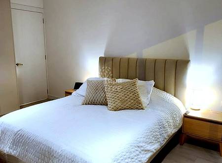 Bedroom with queen bed, upholstered headboard, white linens, two beige pillows, wooden nightstand with lamp, cream walls, and recessed lighting.