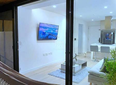 Interior view through large black-framed glass doors showing white walls, brick ceiling, mounted TV, and modern kitchen with recessed lighting