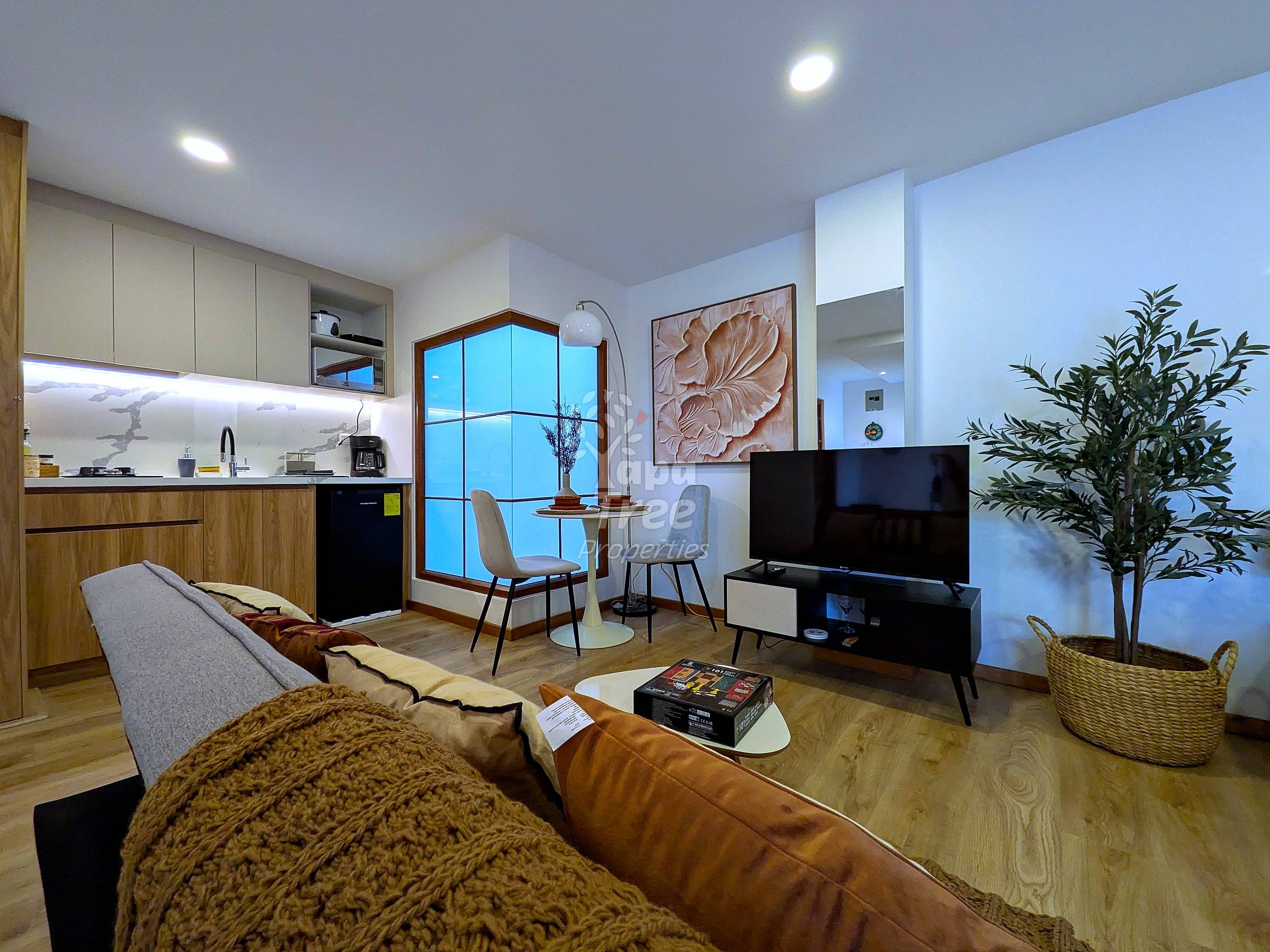 Open-plan suite with wood cabinetry kitchen, brown leather sofa, flat-screen TV on black stand, dining table by window, and potted plant on wooden flooring.
