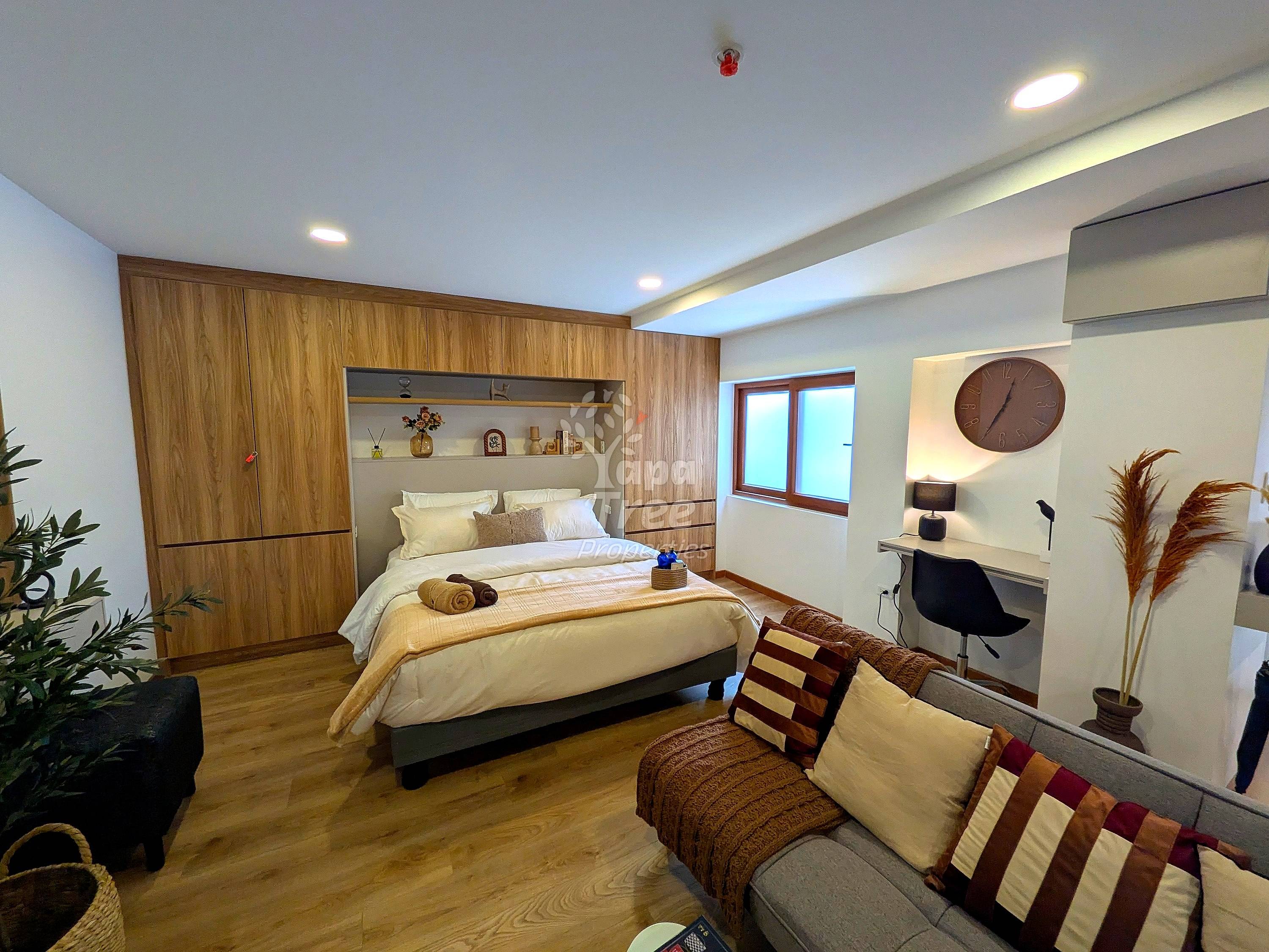 Master bedroom with queen bed, built-in wood cabinetry, cream bedding, seating area with sofa, wooden flooring, recessed ceiling lights, and work desk.