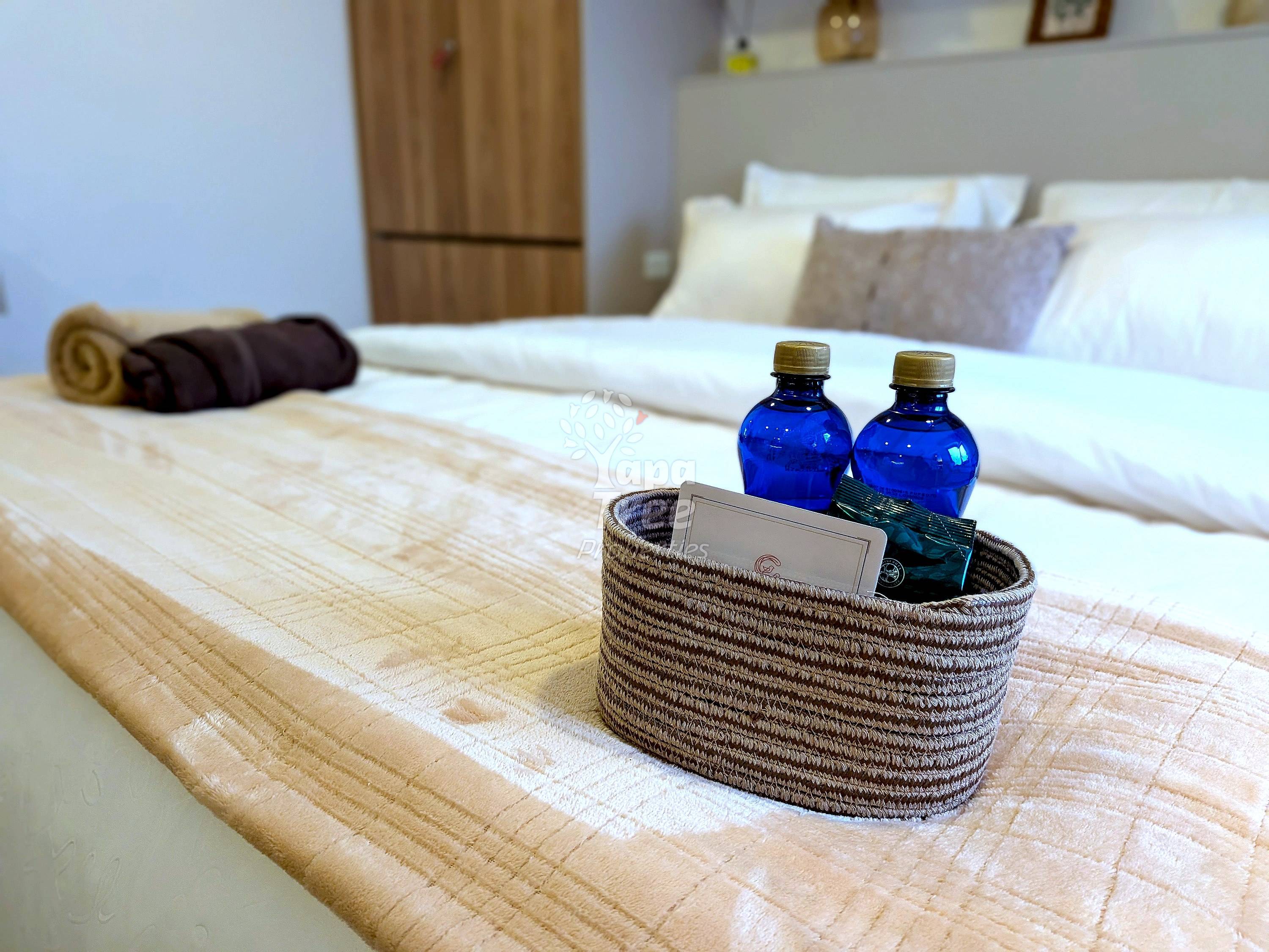 Bedroom with wooden bed frame, white pillows, woven basket with blue bottles, brown rolled towel, and wood wardrobe.
