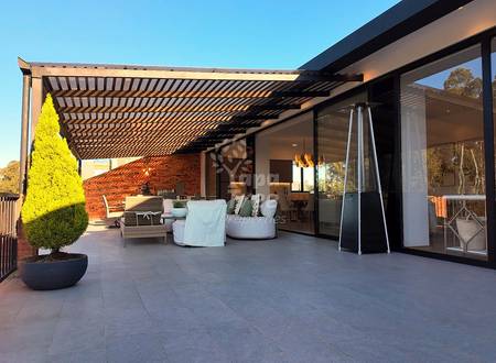 Covered terrace with slatted pergola, grey tile flooring, modern seating area, large sliding glass doors, and brick wall under clear blue sky.