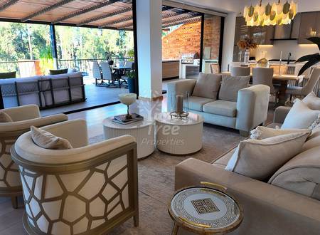 Open-plan living room with cream upholstered seating, round side tables, pendant lighting, brick accent wall, and glass doors opening to terrace with mountain views.