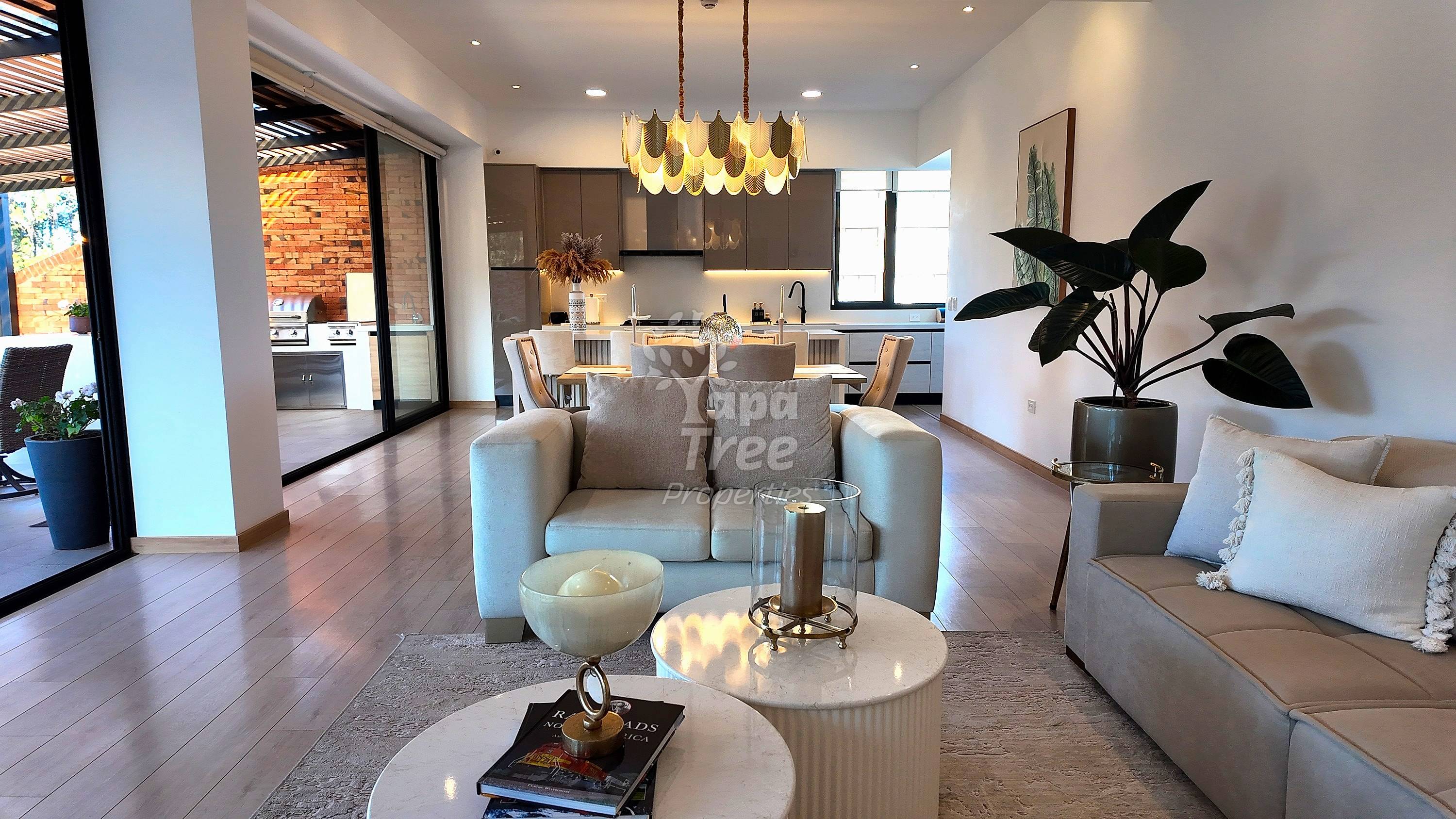 Open-plan departamento with gray upholstered seating, round white tables, pendant lighting, dark kitchen with brick accent wall, and potted plant near windows.