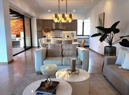 Open-plan departamento with gray upholstered seating, round white tables, pendant lighting, dark kitchen with brick accent wall, and potted plant near windows.