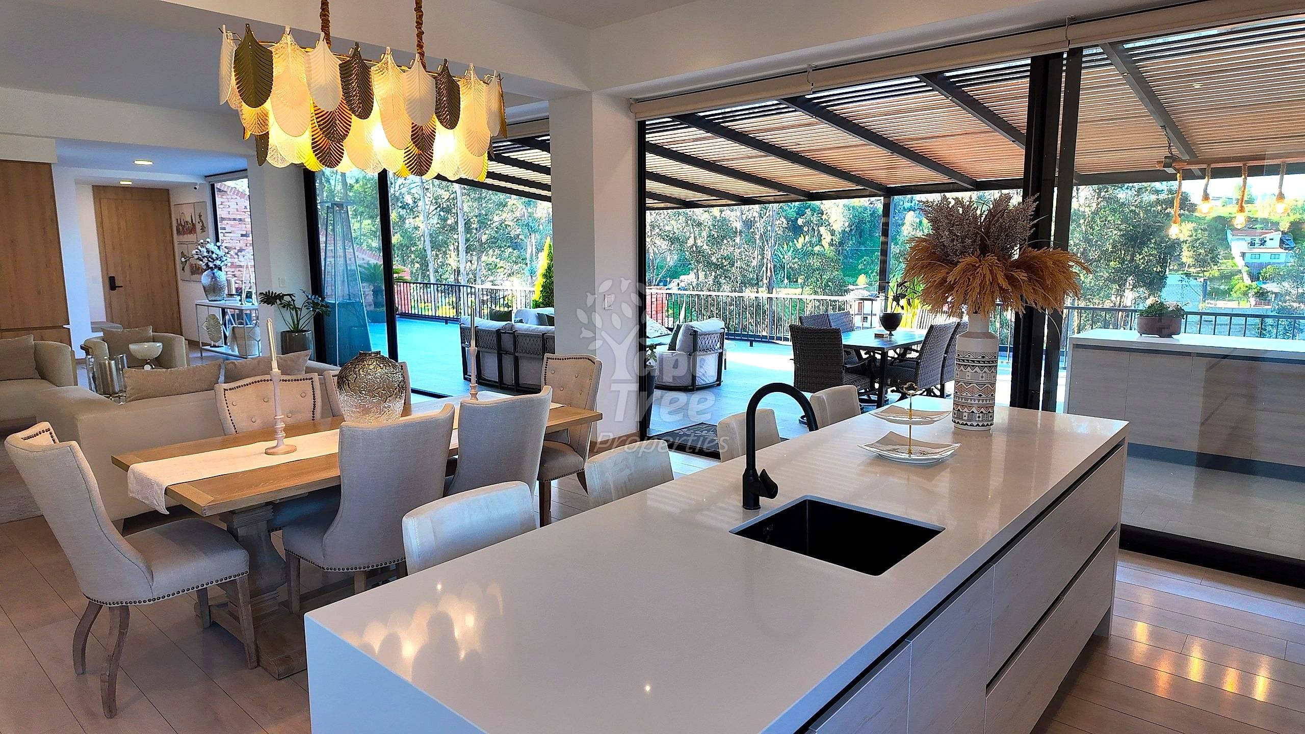 Cocina y comedor de planta abierta con isla de cuarzo blanco, mesa de madera clara, sillas tapizadas, iluminación colgante, y ventanales que dan a terraza y piscina.