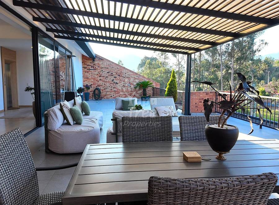 Covered terrace with dining table, woven chairs, lounge seating, slatted pergola roof, and valley views through metal railing.