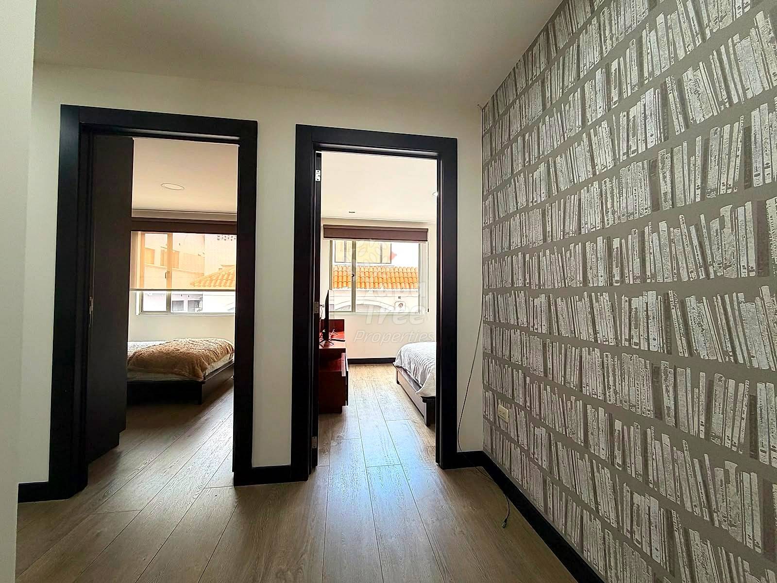 Hallway with wood flooring, black-framed glass doors opening to two bedrooms, decorative brick wall on right, windows with city views.