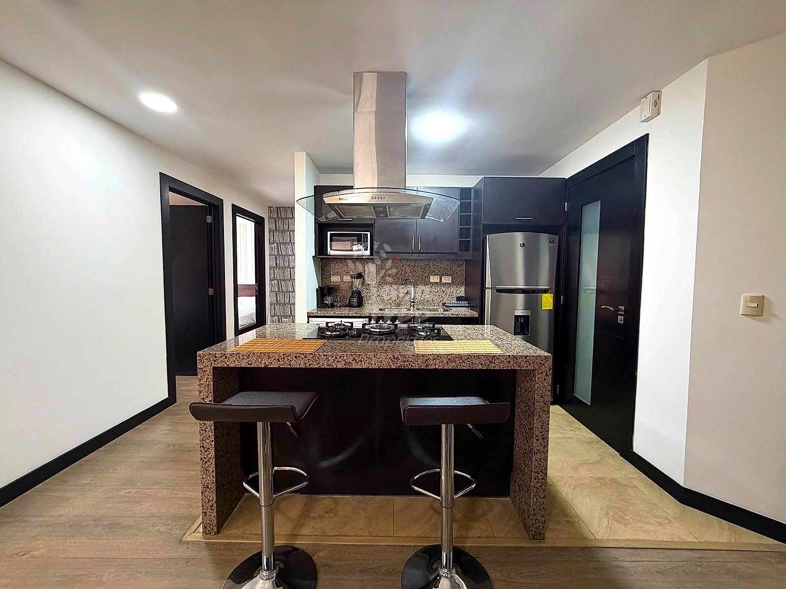 Modern kitchen with dark cabinetry, granite island with two bar stools, stainless steel appliances, gas cooktop, and recessed lighting throughout.