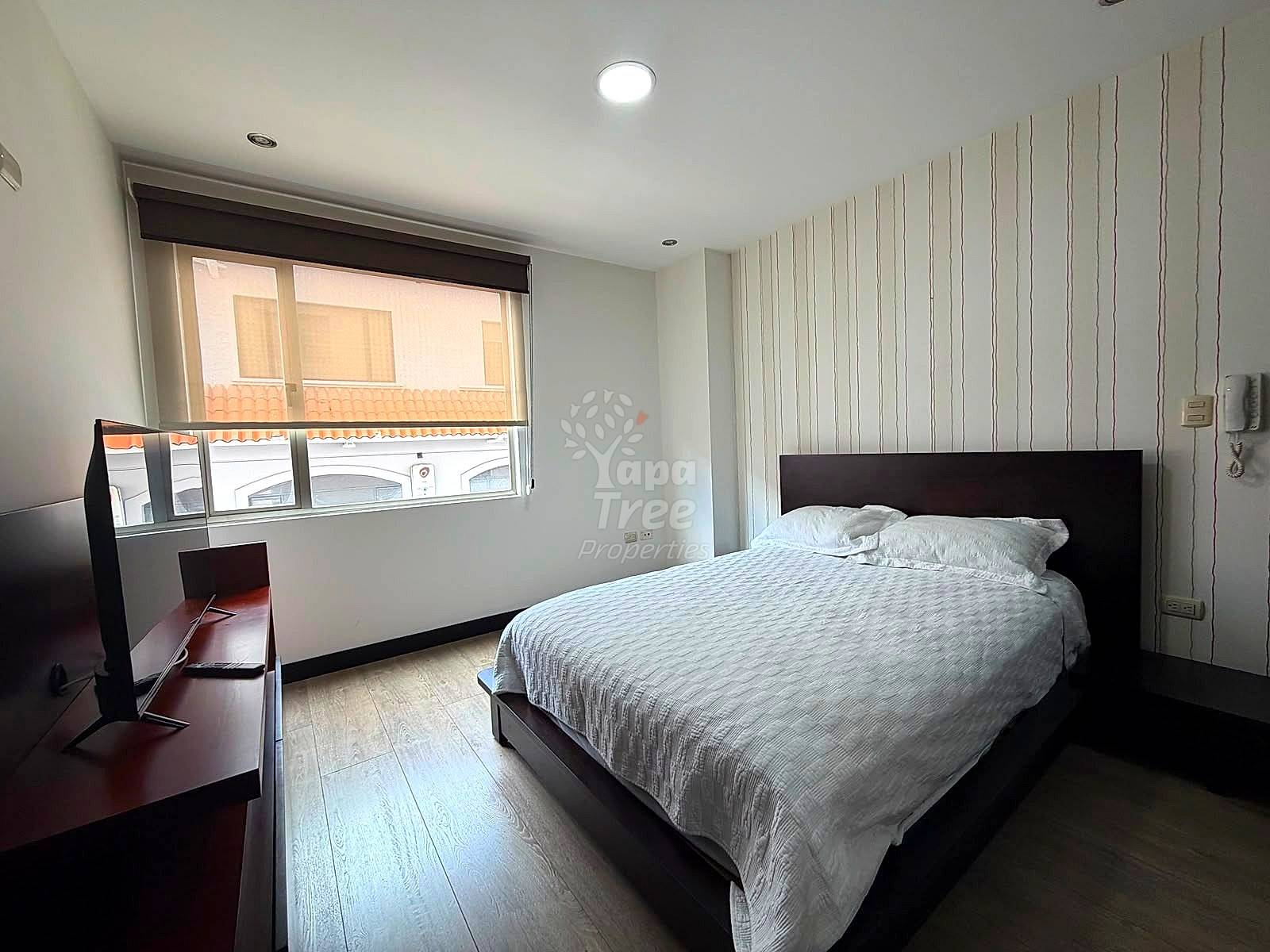 Bedroom with queen bed, black headboard, dark wood desk, vertical striped accent wall, recessed ceiling light, and street-view window with blinds.