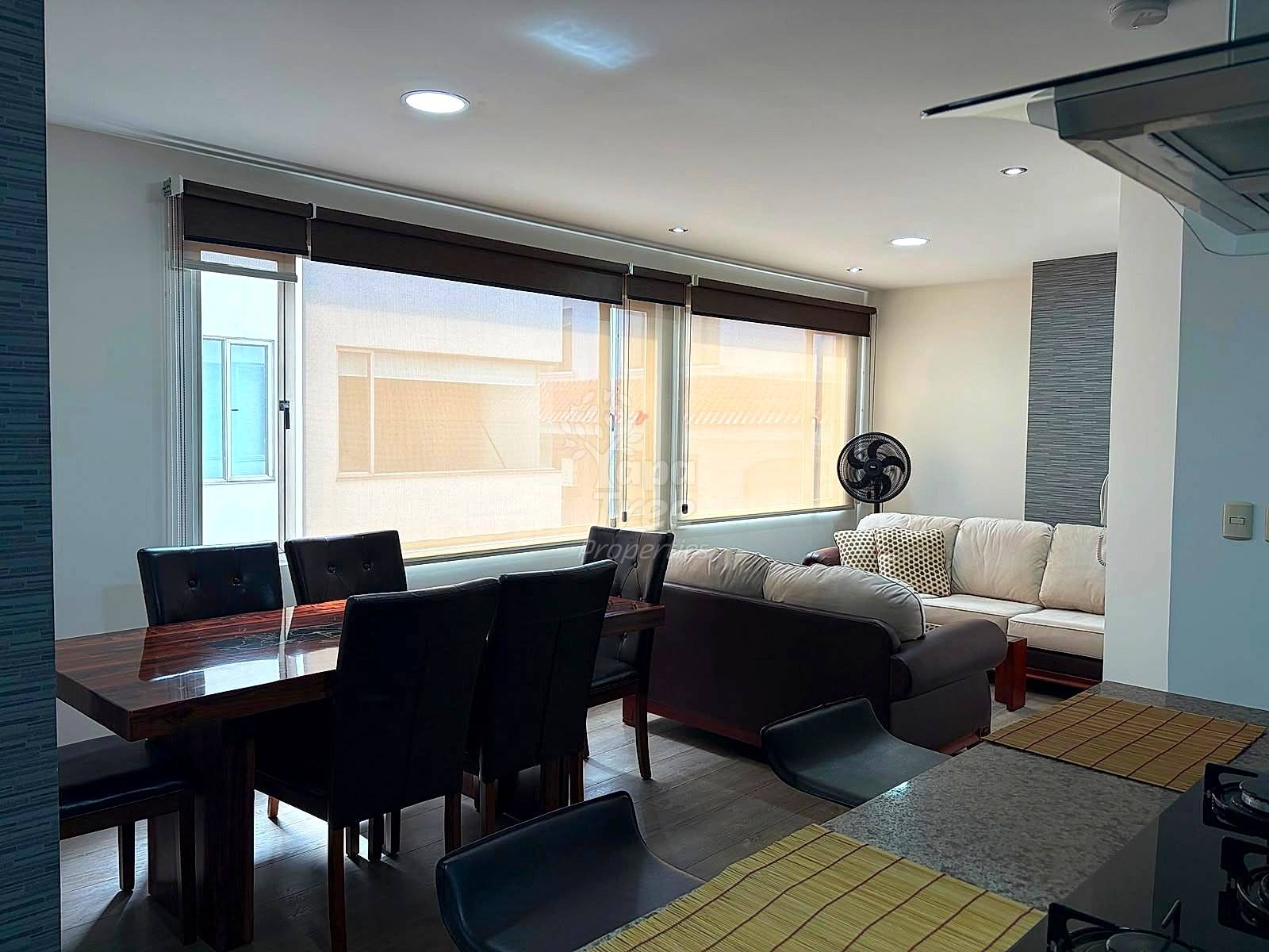 Open-plan living and dining area with black dining table, cream sofa, large windows with black shades, recessed ceiling lights, and tile flooring.