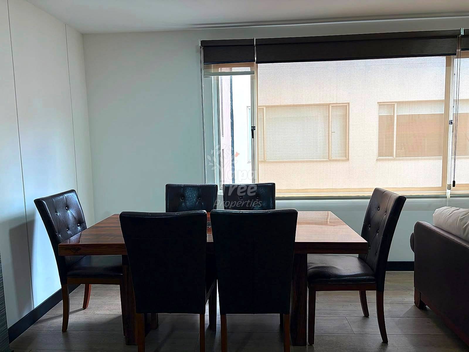 Dining room with wooden table, four black leather chairs, large window with black blinds, light walls, and partial sofa visible.