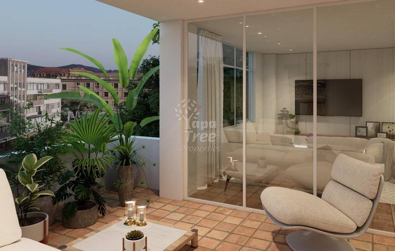 Modern balcony with terra cotta tile floor, potted green plants, beige armchair, glass sliding doors opening to interior room with TV and seating.