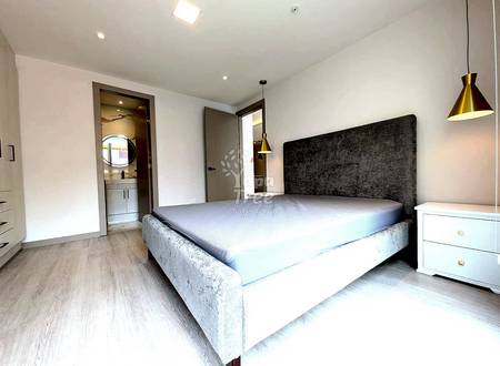 Modern bedroom with queen bed, gray upholstered headboard, light wood flooring, white nightstand, pendant lights, and en-suite bathroom visible through open door.