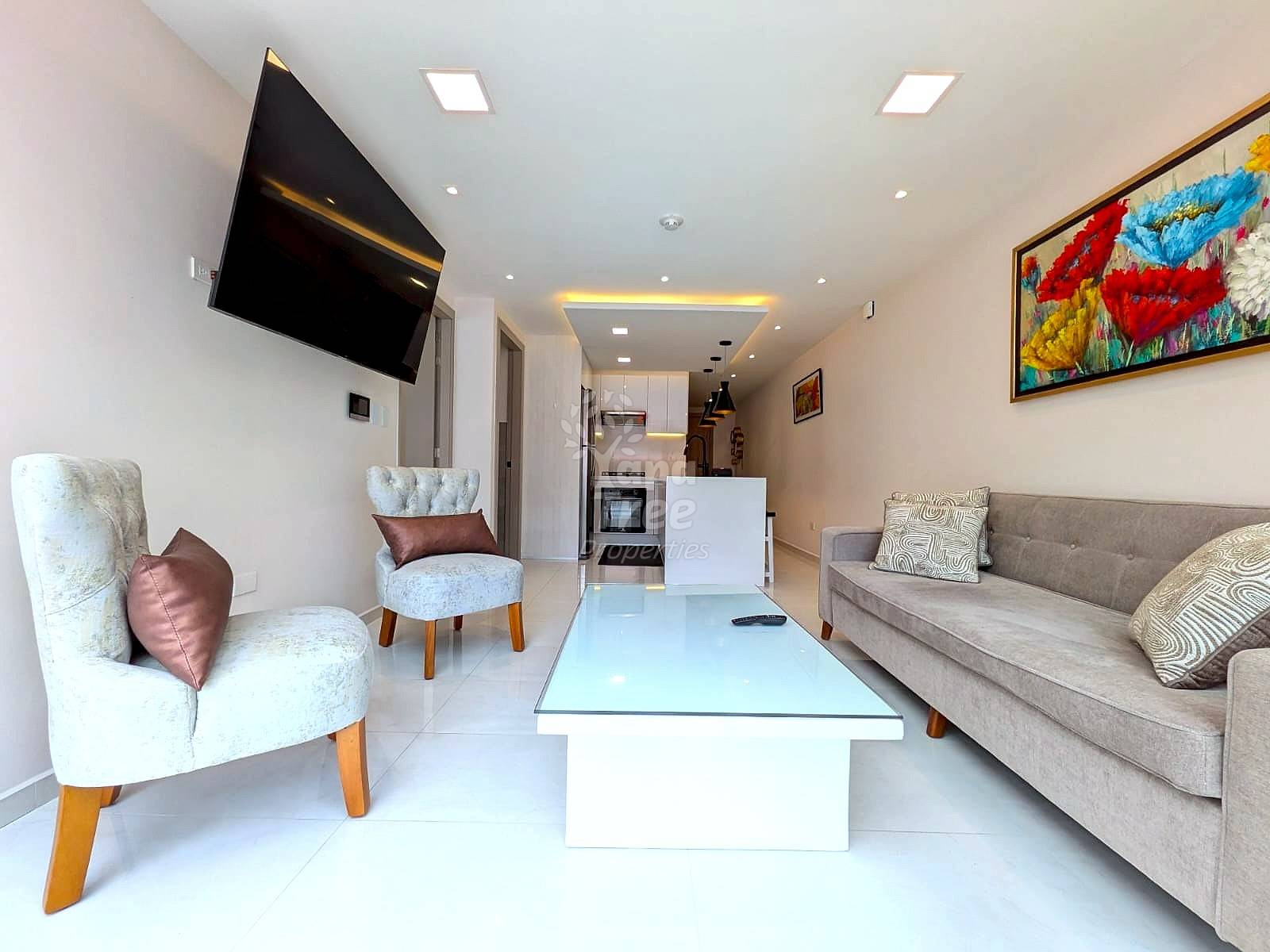 Open-plan living room with light gray sofa, cream accent chair, glass coffee table, wall-mounted TV, recessed ceiling lights, and kitchen visible in background.