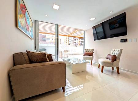 Modern living room with beige tile floors, brown sofa, cream accent chairs, glass coffee table, wall-mounted TV, and sliding glass doors opening to a terrace.