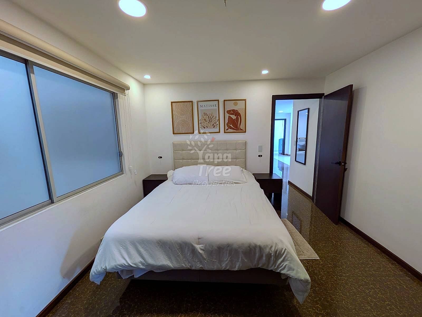 A modern bedroom with white walls, recessed lighting, and a bed with a cream tufted headboard.
