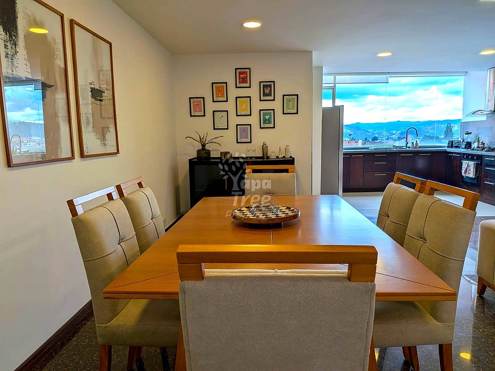 A dining room with a wooden table and upholstered chairs, next to a kitchen with a view.