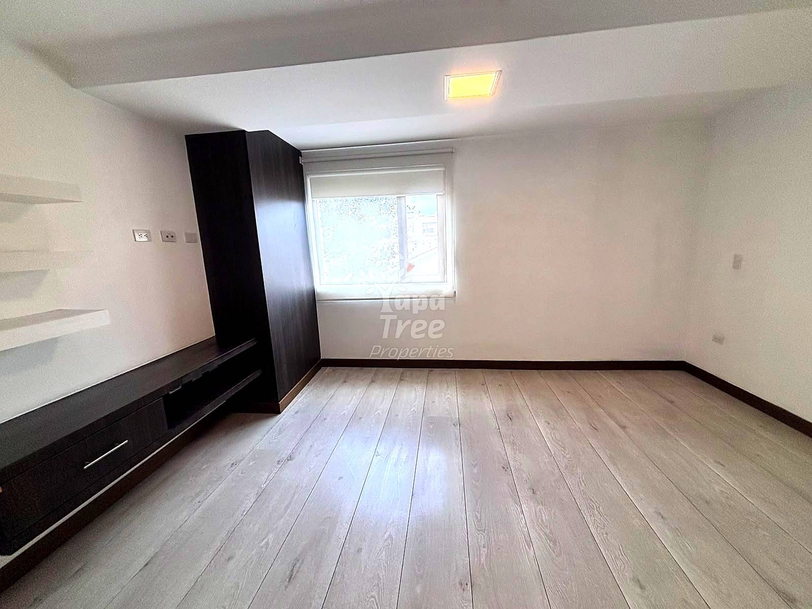 Empty living room with light wood plank flooring, white walls, black built-in cabinet, window with street view, and recessed ceiling light.