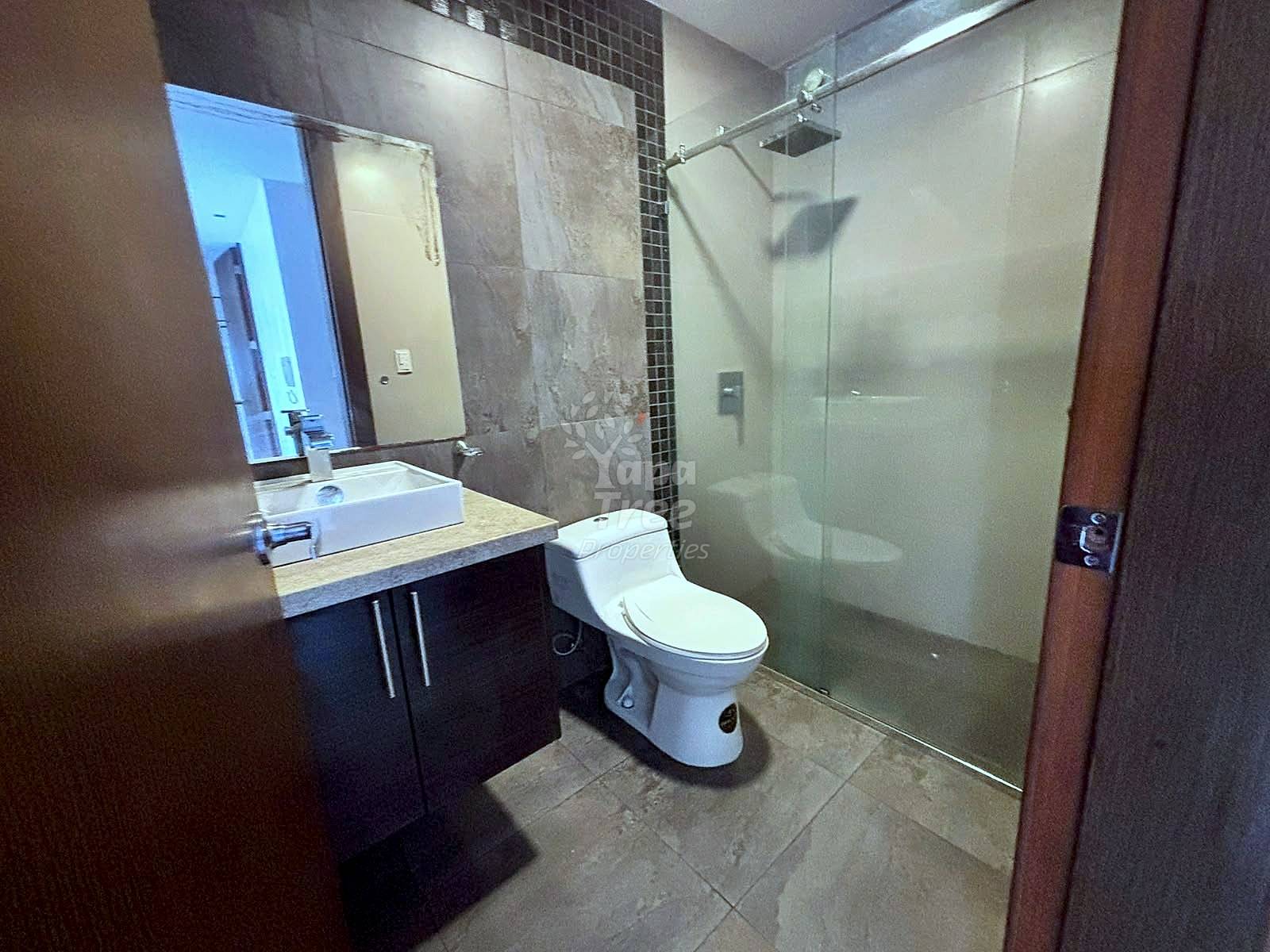 Bathroom with white rectangular sink on dark vanity, toilet, glass shower enclosure, gray tile walls, and large mirror with lighting.