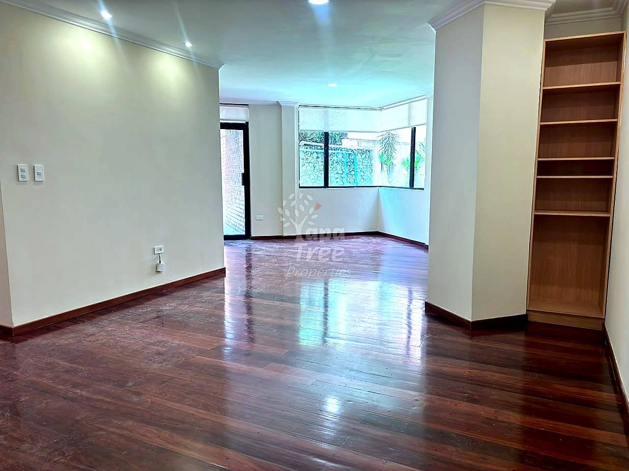 Open living room with dark wood flooring, white painted walls, recessed ceiling lights, large windows with garden views, and wooden built-in shelving on right side.