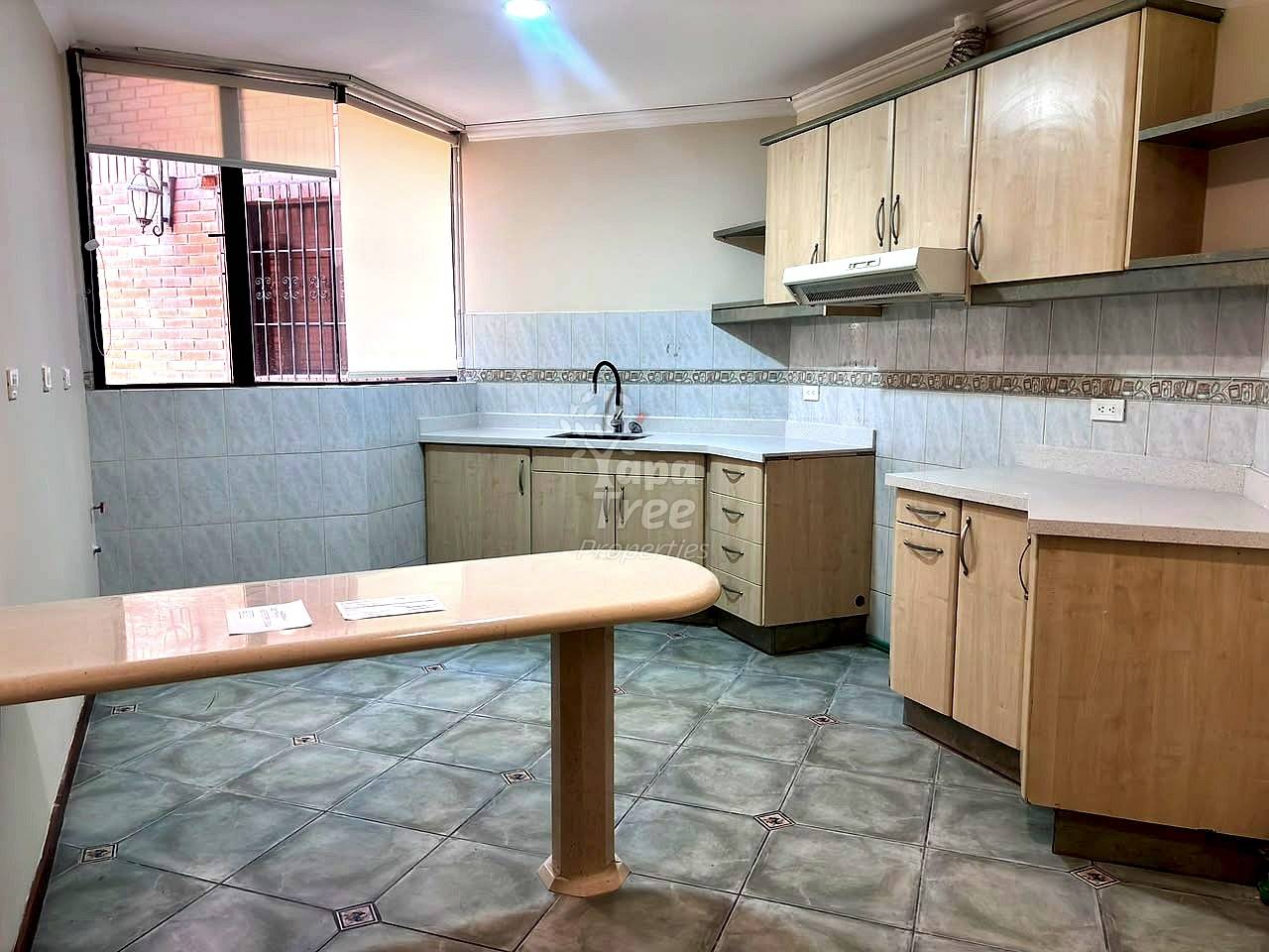 Kitchen with light wood cabinets, tiled countertops, central table, window with security bars, and gray tile flooring.
