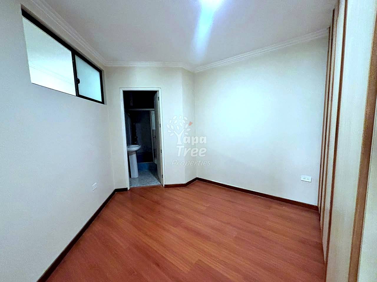 Empty bedroom with laminate wood flooring, cream walls, window with black frame, and open doorway to bathroom.