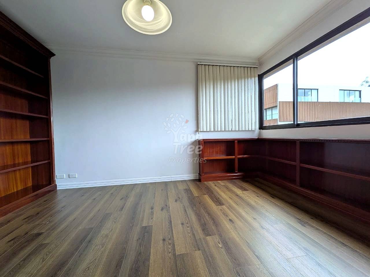 Empty room with wood laminate flooring, white walls, ceiling light fixture, large window with vertical blinds, and wooden built-in shelving units on two walls.